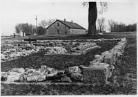 Stone foundation ruins in the foreground of a black-and-white image, with a tree and building in the background.